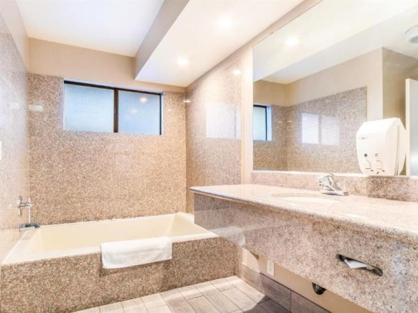 This image shows a clean, modern bathroom with a bathtub, a large countertop with a sink, and a mirror above. The walls are tiled, and lighting is ample.
