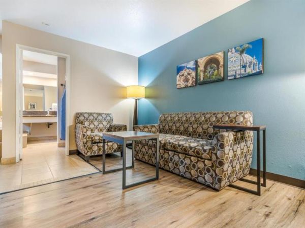 The image shows a modern living area with a patterned couch, two matching chairs, a coffee table, and wall art; a bathroom is visible in the background.