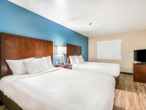 A hotel room with two double beds, a nightstand with a lamp, a TV on a dresser, wooden floors, and a window on the far wall.