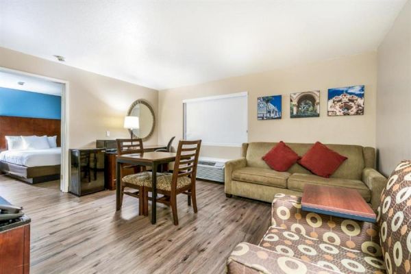 This image shows a hotel room with a bed, a seating area, a dining table with chairs, and wall décor. The room has wooden flooring.