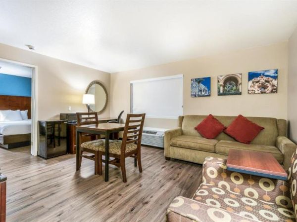 This image shows a hotel room with a bed, a seating area, a dining table with chairs, and wall décor. The room has wooden flooring.