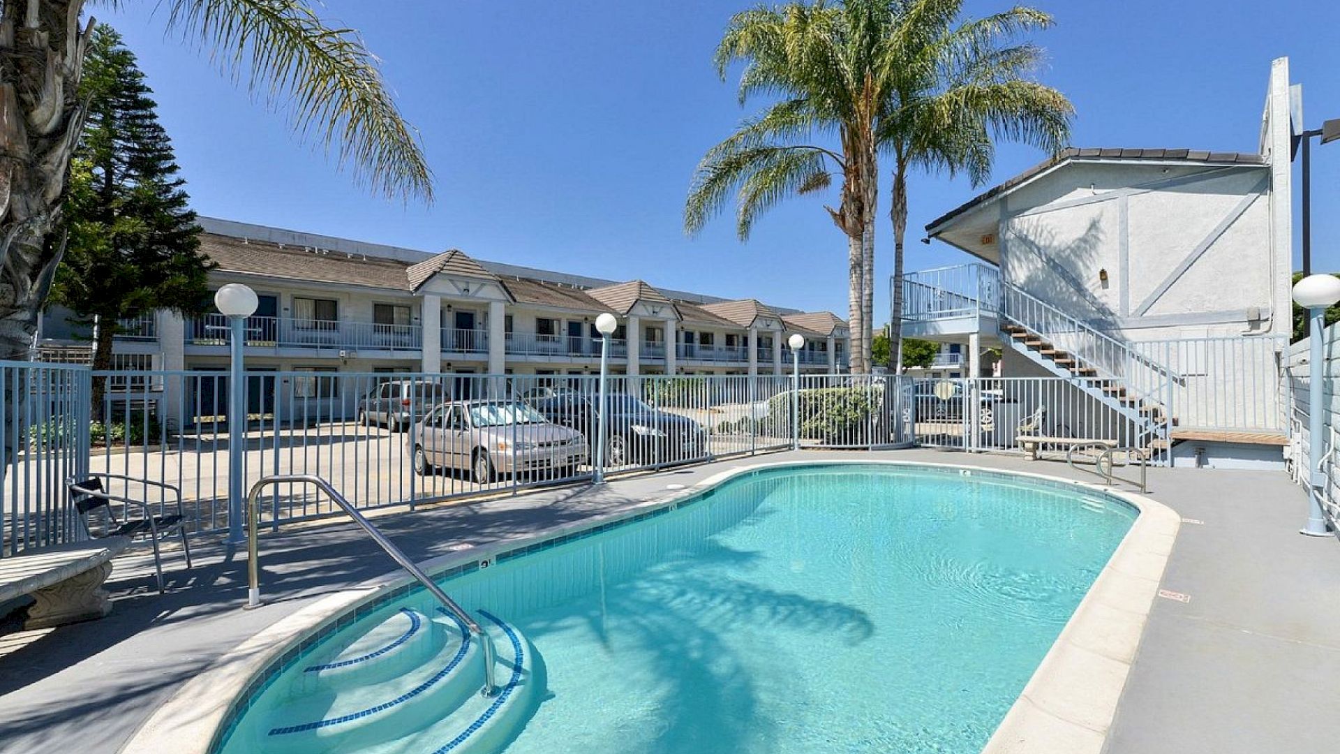 An outdoor pool surrounded by a fence with palm trees, stairs, and a building in the background under a clear blue sky.