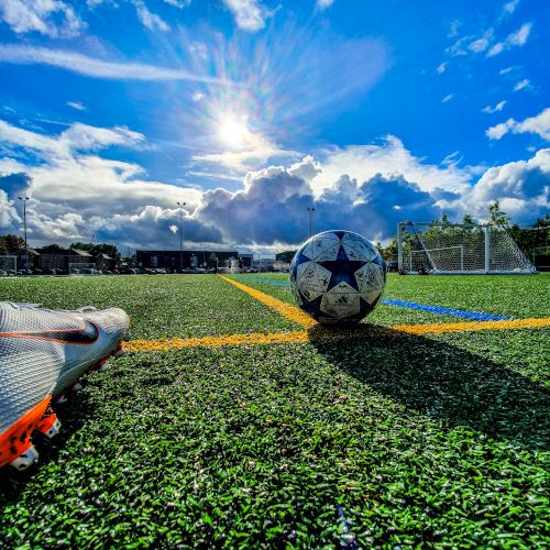 A soccer field with a ball, cleats, and goals in the background under a bright, partly cloudy sky.