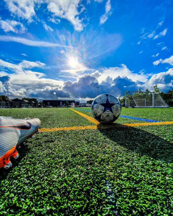 A soccer field with a ball, cleats, and goals in the background under a bright, partly cloudy sky.