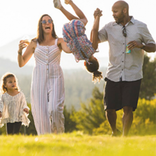 A family of four enjoys outdoor fun, with the parents swinging one child while another child walks beside them on a sunny day.