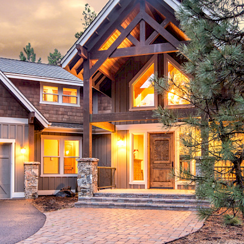 The image shows a large, modern house with a wooden exterior, multiple gables, lit windows, and a driveway, surrounded by trees at dusk.