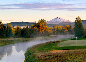 A golf course by a river with snow-capped mountains in the background, surrounded by trees under a colorful sky at sunrise or sunset.
