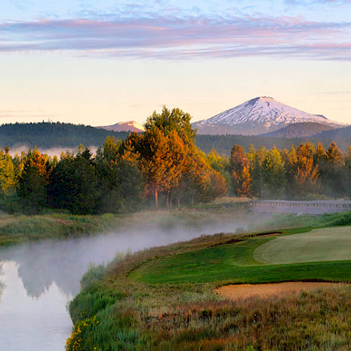 A golf course by a river with snow-capped mountains in the background, surrounded by trees under a colorful sky at sunrise or sunset.
