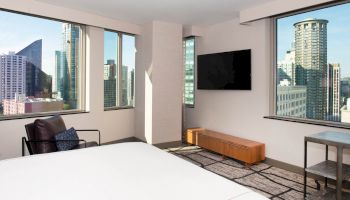 A modern room with a large bed, TV, leather chair, and cityscape views through three large windows, providing a bright and airy atmosphere.