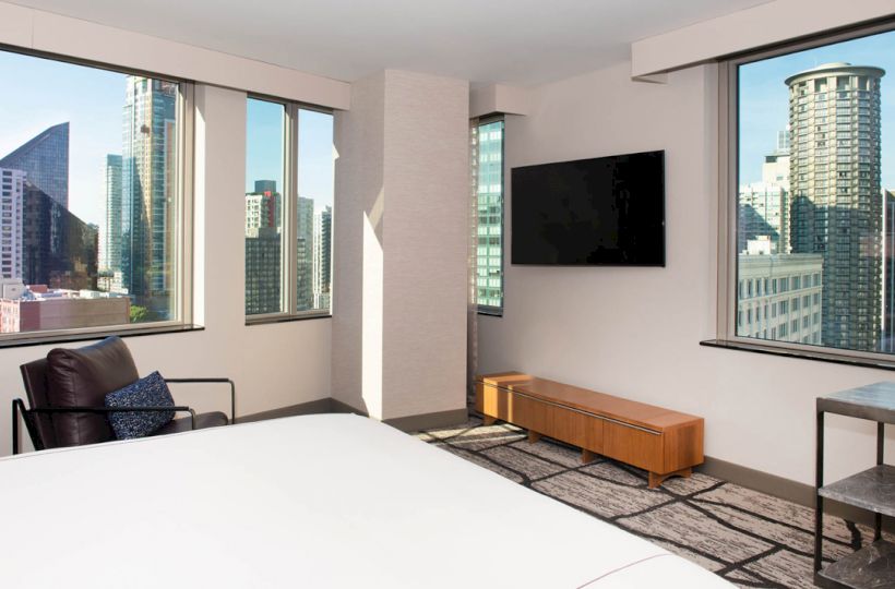 A modern room with a large bed, TV, leather chair, and cityscape views through three large windows, providing a bright and airy atmosphere.