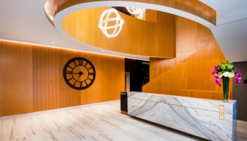 A modern lobby with a marble reception desk, a wooden curved staircase, a wall clock, and decorative lighting fixtures.