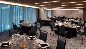 A banquet hall set up with round tables, each adorned with black napkins, glassware, and flower centerpieces, under modern lighting fixtures.