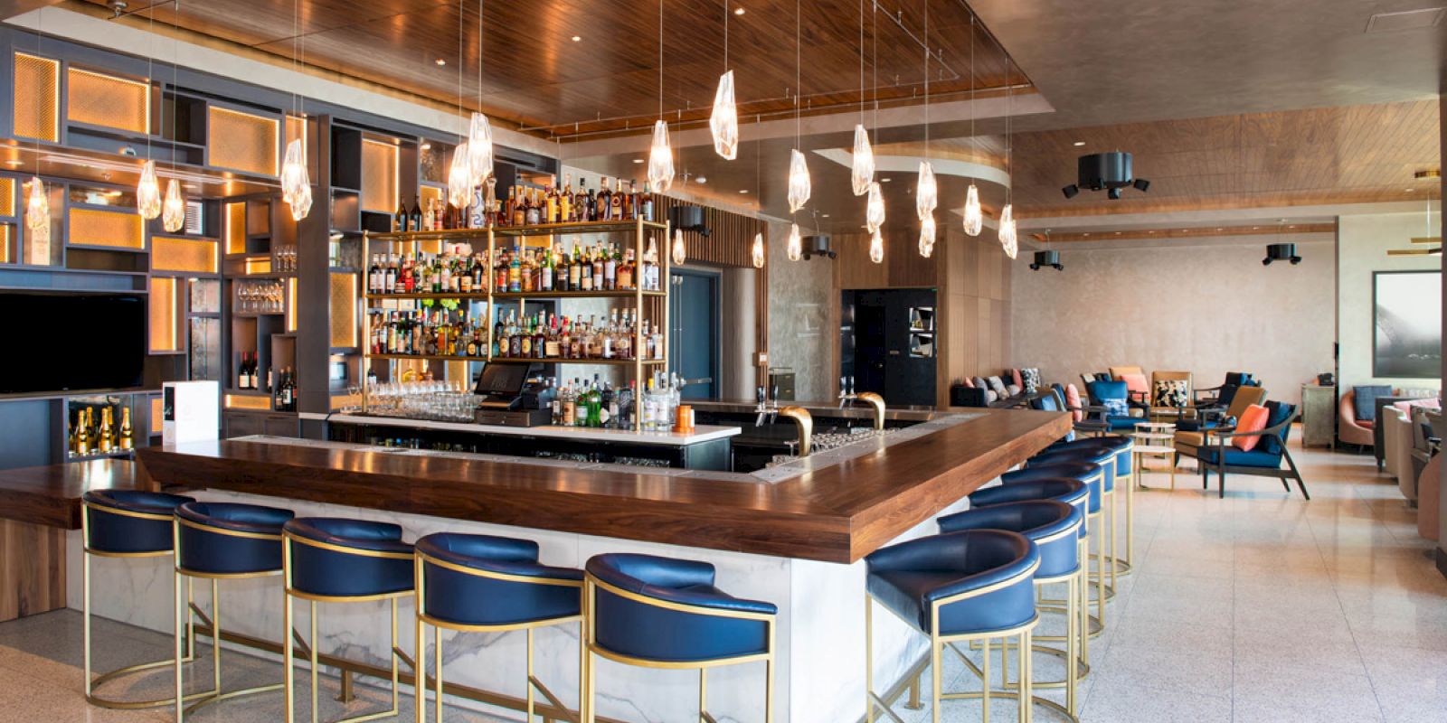 The image shows a modern bar with a well-stocked counter, blue and gold bar stools, overhead lighting fixtures, and a lounge area in the background.