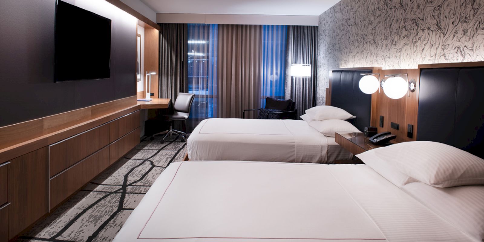 The image shows a modern hotel room with two neatly made beds, a TV on the wall, a desk with a chair, and stylish decor.