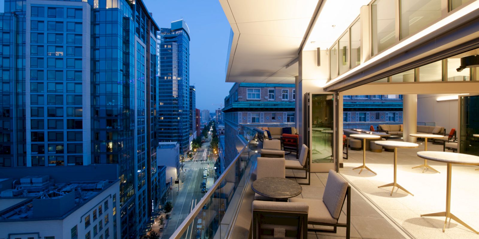 This image shows a modern rooftop terrace with tables and chairs overlooking a city street lined with tall buildings at dusk, with city lights on.