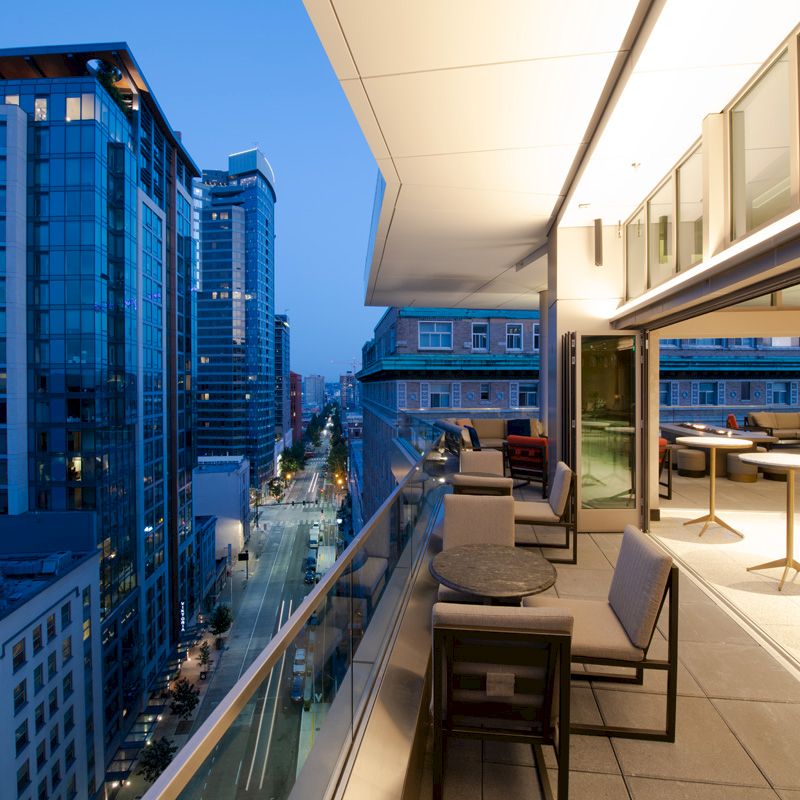 A modern urban balcony with outdoor seating overlooks a city street lined with tall buildings, illuminated by evening lights.