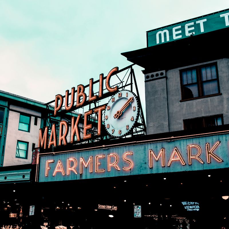 The image shows the entrance to a public market with neon signs reading "Public Market" and "Farmers Market," along with a clock displaying the time.