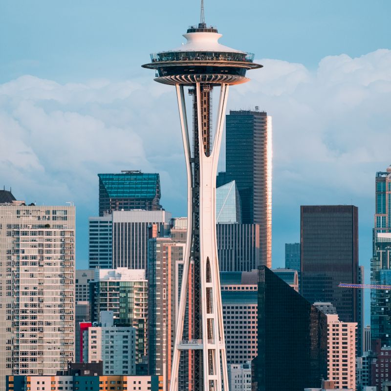 The image shows the Space Needle skyscraper surrounded by other buildings against a cloudy sky in what looks like a bustling cityscape setting.