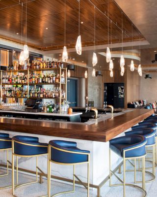 A modern bar with a stocked liquor shelf, stylish pendant lights, and blue cushioned stools. The room has a contemporary design with seating areas.