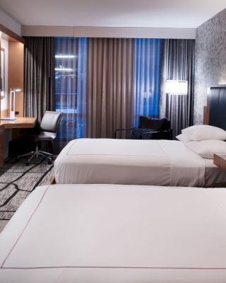 The image shows a modern hotel room with two beds, a desk, chair, large TV, and stylish decor.