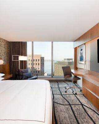 This image shows a modern hotel room with a large bed, a wall-mounted TV, a desk with a chair, and a city view through ceiling-to-floor windows.