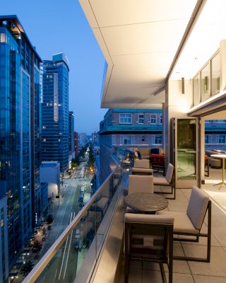 A modern balcony view with outdoor seating, overlooking a cityscape of tall buildings and a lit street during twilight hours.
