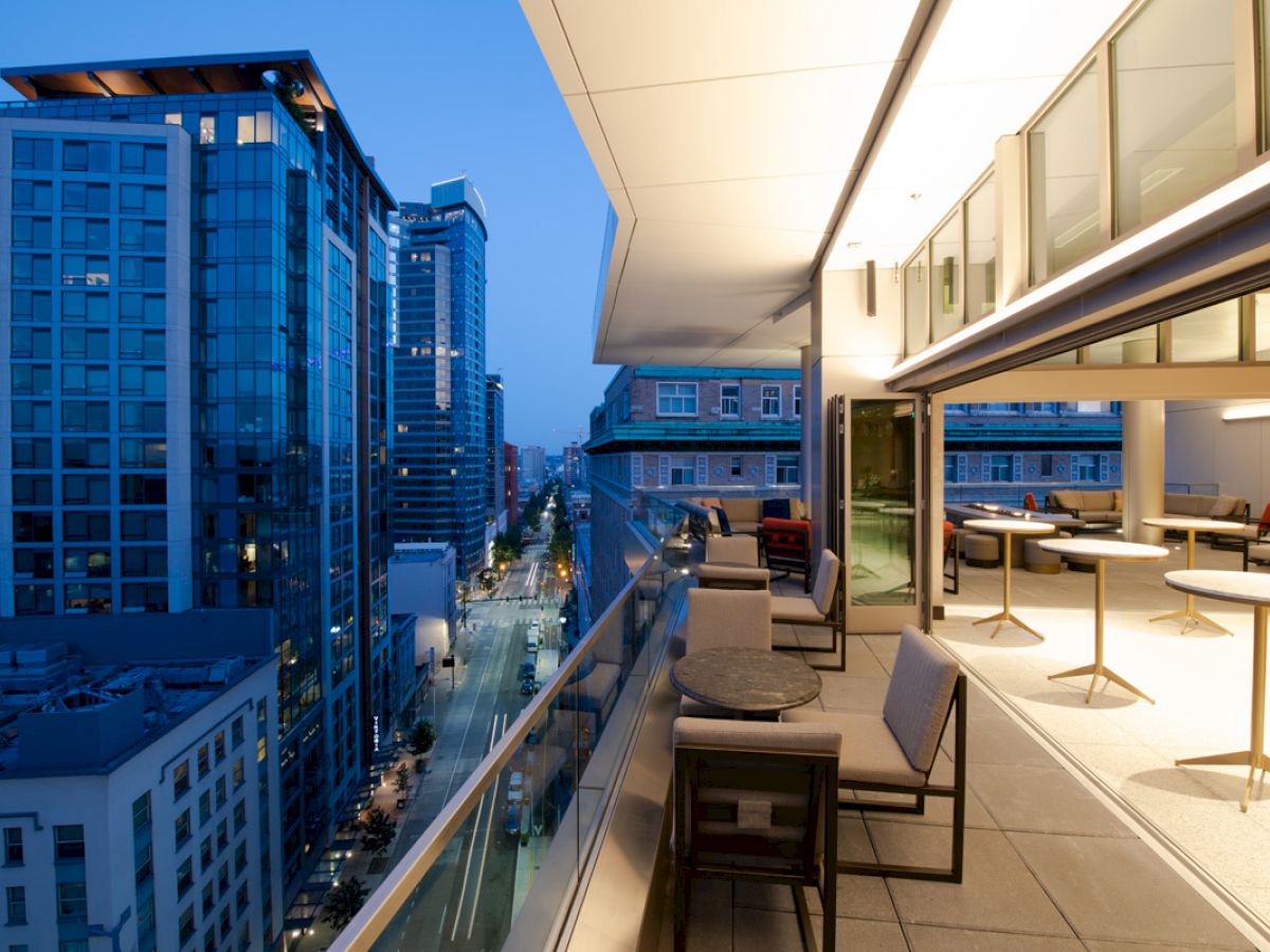 An urban balcony setup with tables and chairs overlooks a city street with tall buildings at dusk, lights illuminating the scene.