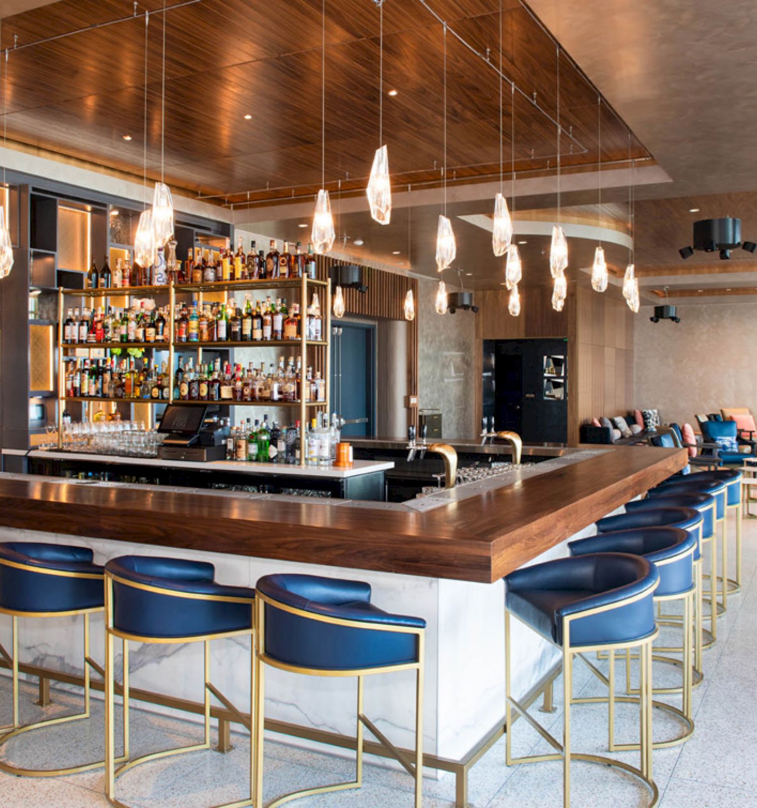 The image shows a modern bar with a wooden counter, high chairs, hanging lights, and a fully stocked drink display behind.