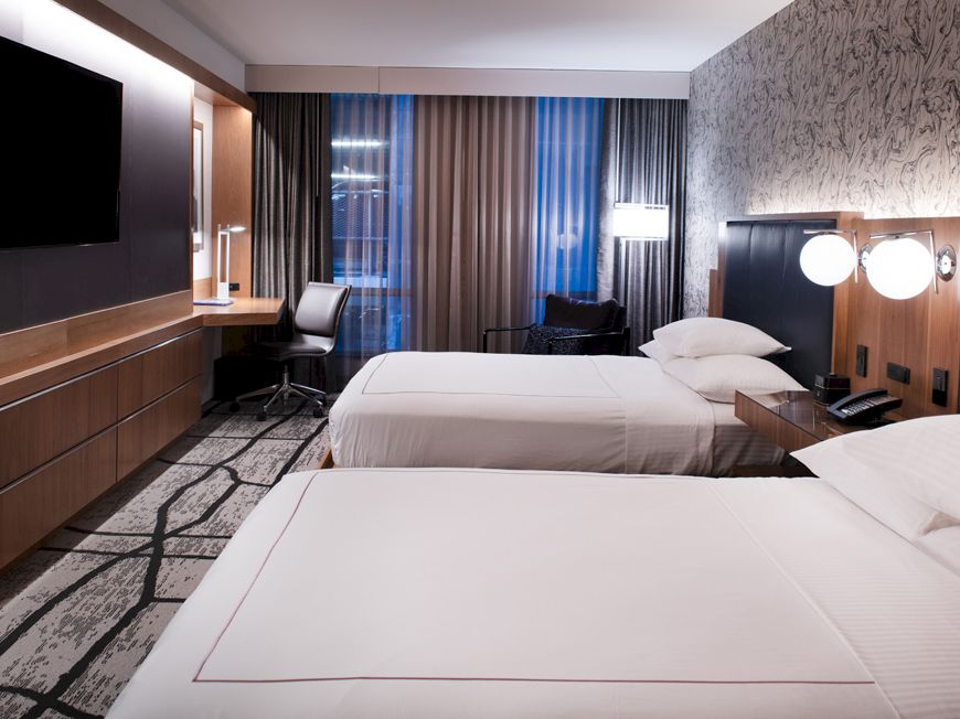A modern hotel room features two neatly made beds, a wall-mounted TV, a desk with a chair, and floor-to-ceiling windows with curtains.