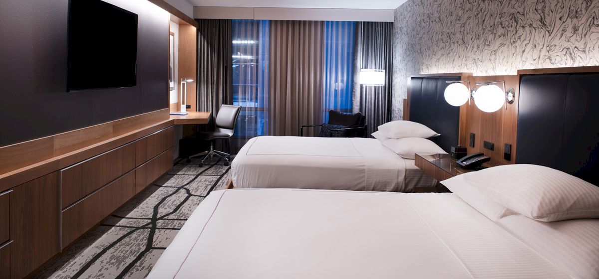 A modern hotel room features two neatly made beds, a wall-mounted TV, a desk with a chair, and floor-to-ceiling windows with curtains.
