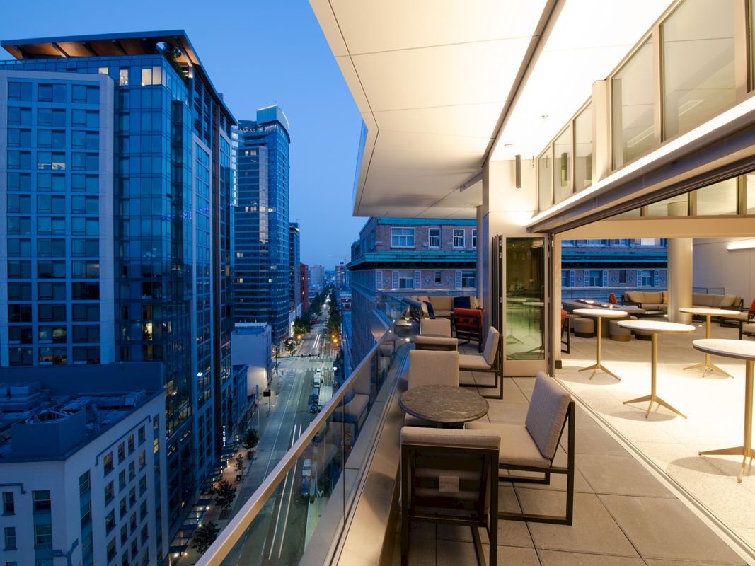 The image shows a view from a well-lit, furnished balcony overlooking a cityscape with several modern high-rise buildings at dusk.