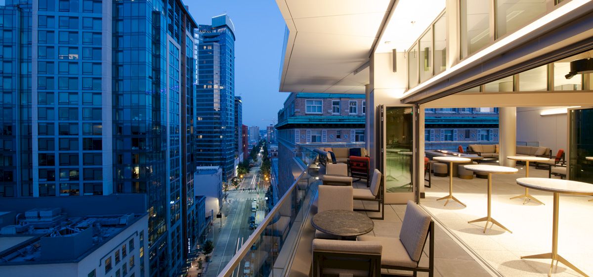 The image shows a view from a well-lit, furnished balcony overlooking a cityscape with several modern high-rise buildings at dusk.