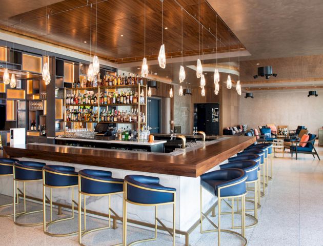 This image shows a modern bar with a large selection of bottles, stylish barstools, and ambient lighting in a spacious, well-designed interior.