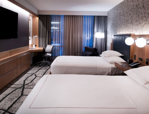 The image shows a modern hotel room with two neatly made beds, a TV, a desk with a chair, and contemporary lighting and decor.