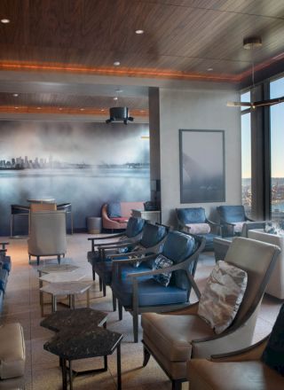 The image shows a modern lounge area with numerous cushioned chairs, a wall art display, large windows with a coastal view, and a Ferris wheel in sight.