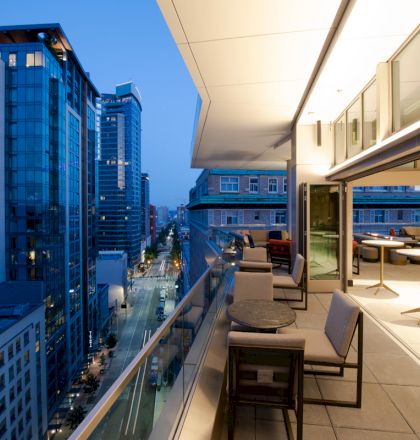 A modern cityscape viewed from a stylish balcony with seating, overlooking a street lined with tall buildings in the evening light.