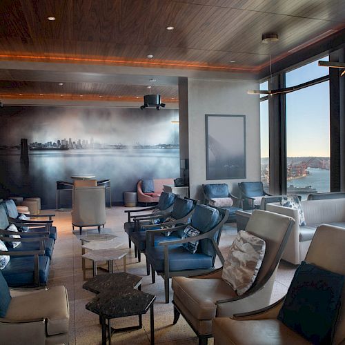 A modern, comfortable lounge area with multiple seating arrangements, large windows offering a scenic view of a waterfront and a skyline mural.