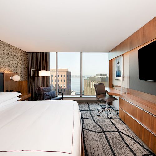 A modern hotel room features a large bed, a workspace with a chair, a flat-screen TV, and a window with a cityscape view.