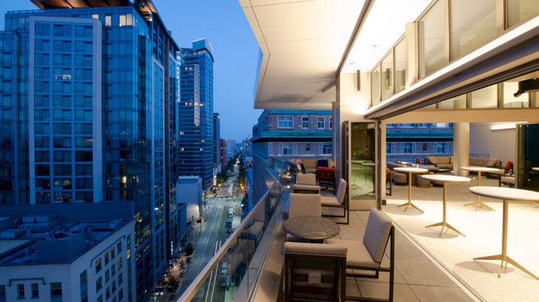 A modern rooftop balcony with seating overlooks a city street lined with tall buildings, illuminated in the evening light.