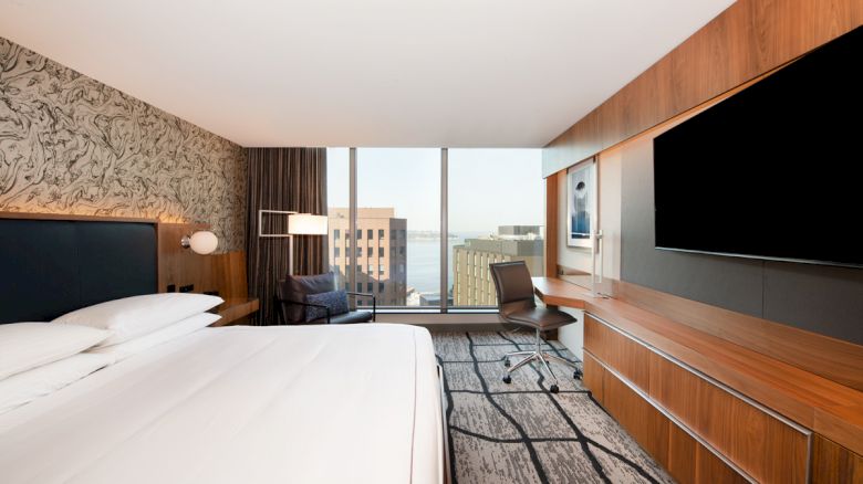 A modern hotel room with a king-sized bed, a large TV, a desk, a chair, and a view of buildings and the water through the window ending the sentence.