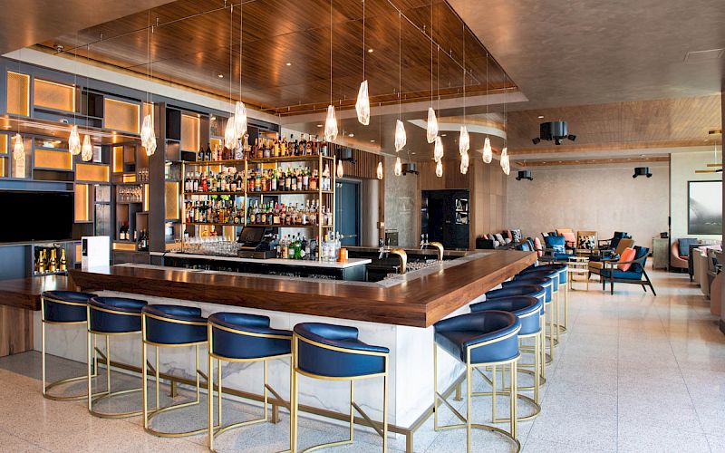 The image features a modern bar with a large countertop, barstools, and a well-stocked collection of bottles behind it. The interior has stylish lighting.