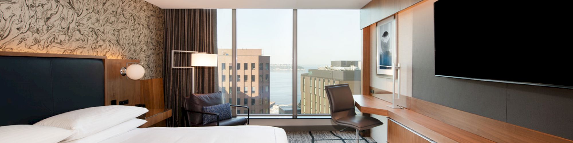 A modern hotel room with a large bed, a desk, a chair, a flat-screen TV, and a window with a city view. The room has sleek and contemporary decor.