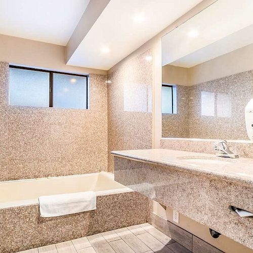 A modern bathroom with a rectangular bathtub, a large vanity mirror, and a granite countertop, with a towel hanging by the bathtub.