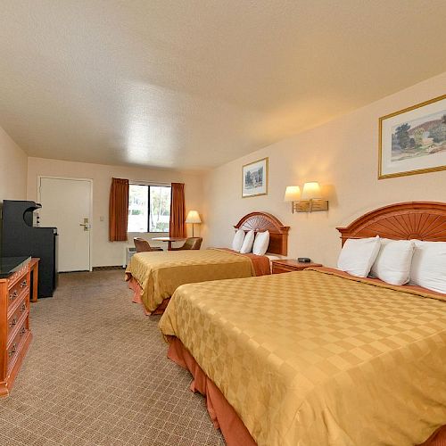 A hotel room with two double beds, a TV, a desk, and a window with brown curtains. It has beige walls and framed artwork.