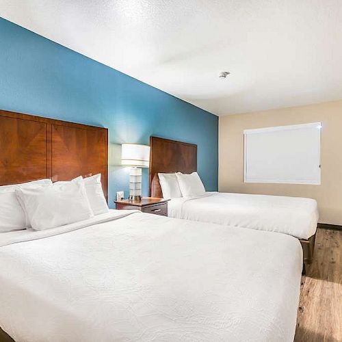 A hotel room with two double beds, a nightstand with a lamp, a TV on a dresser, and a window; blue accent wall behind the beds.