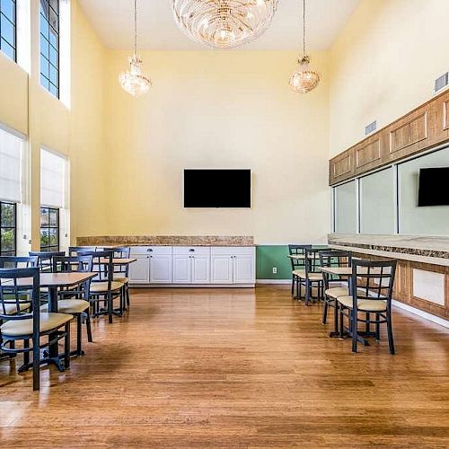 The image shows a dining area with several tables and chairs, a wall-mounted TV, large windows, mirrors, white cabinets, and wooden flooring.