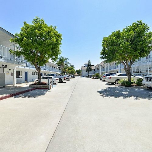 The image shows a courtyard between buildings with walkways, trees, and parked cars on both sides, in a sunny setting.