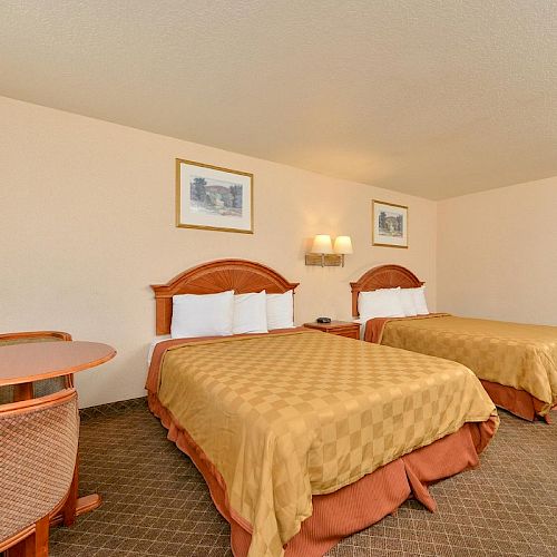 The image shows a hotel room with two double beds, a small table with chairs, framed pictures, and a bathroom visible in the background.