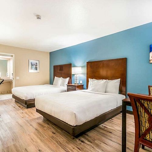 This image shows a hotel room with two double beds, a desk with a chair, and a sitting area, featuring modern decor and hardwood floors.