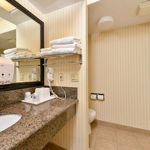 A bathroom with a granite countertop sink, mirror, folded towels, wall-mounted hairdryer, soap dispenser, and a shower stall in the background.